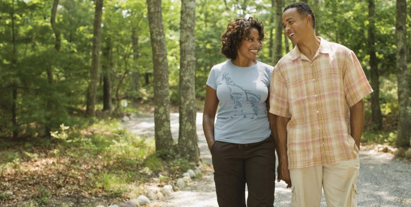 A couple joyfully walks hand-in-hand along a gravel path in a lush, green forest, enjoying each other's company and nature.