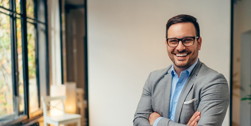 Smiling man in a grey suit stands confidently in a well-lit, modern office space, symbolizing professionalism and approachability.