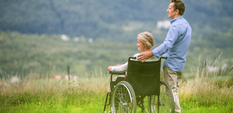 A woman in a wheelchair enjoys a scenic view as a man stands behind her, offering support and companionship in a serene outdoor setting.