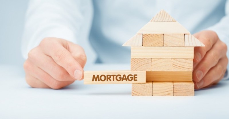 Hand holding a wooden block labeled "MORTGAGE" being inserted into a small wooden house model, symbolizing the concept of home financing.