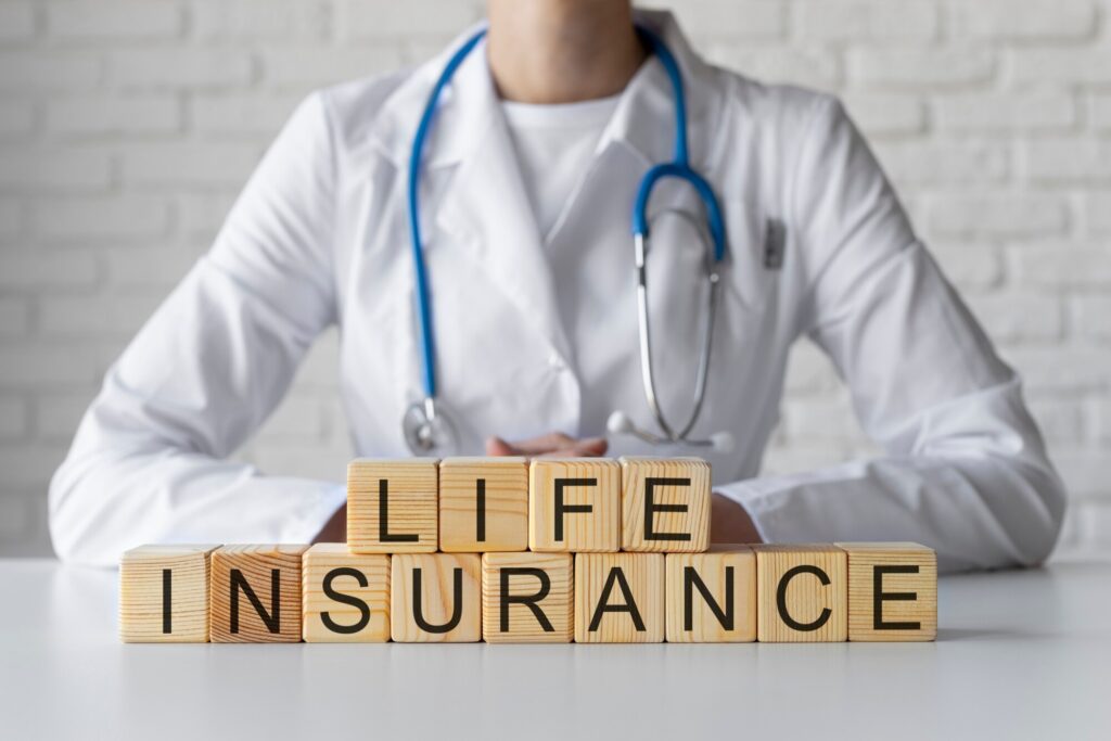A healthcare professional in a lab coat sits behind wooden blocks spelling "LIFE INSURANCE," emphasizing the connection between health and financial planning.