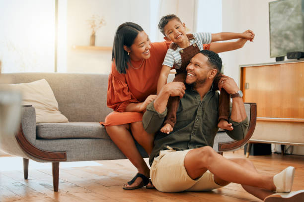 A joyful family scene features a smiling father sitting on the floor with his son on his back, while the mother playfully leans over the couch.